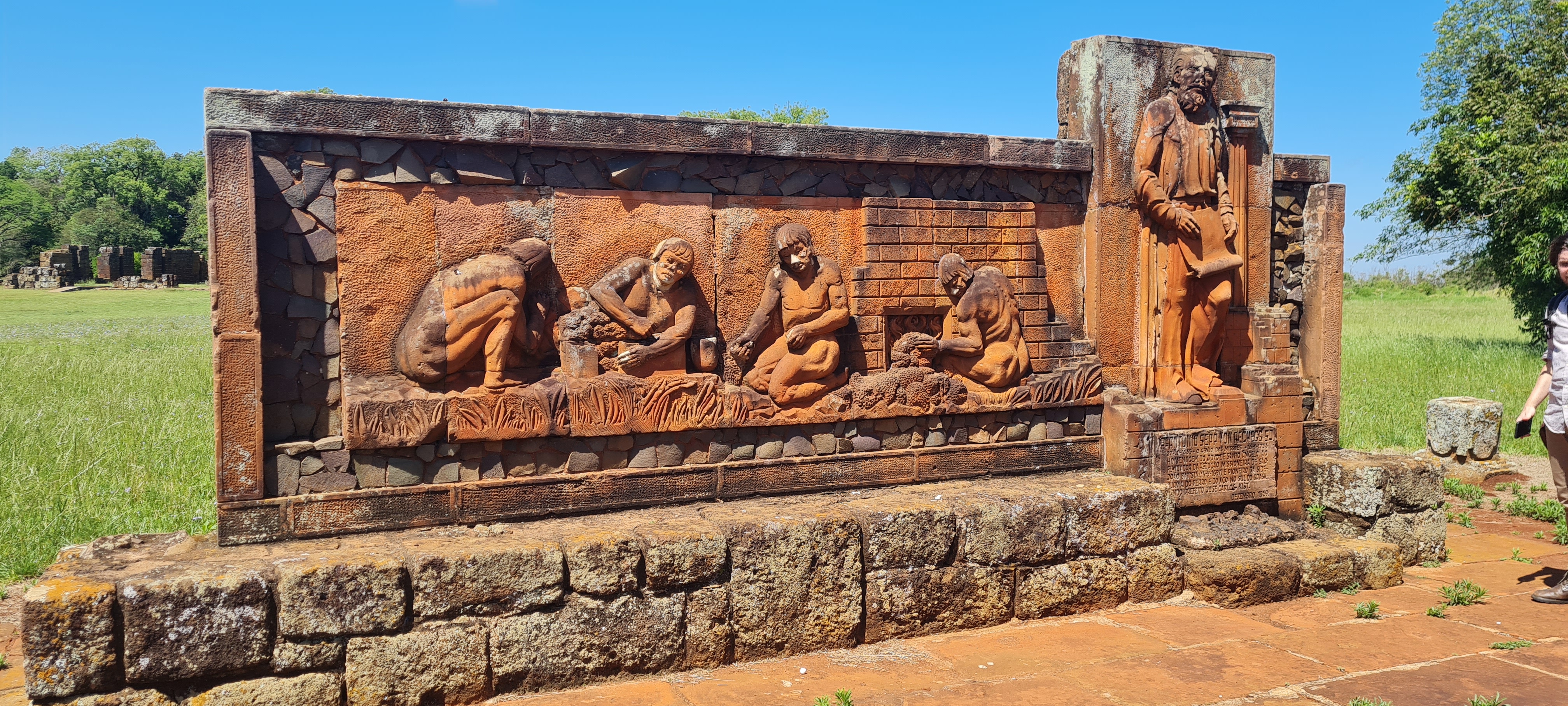 A fotografia colorida mostra uma pedra alaranjada com entalhes de indígenas trabalhando com ferro ao lado de um padre