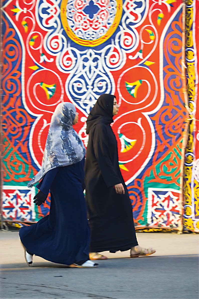 Mulheres de roupas escuras andando em frente de arabescos coloridos