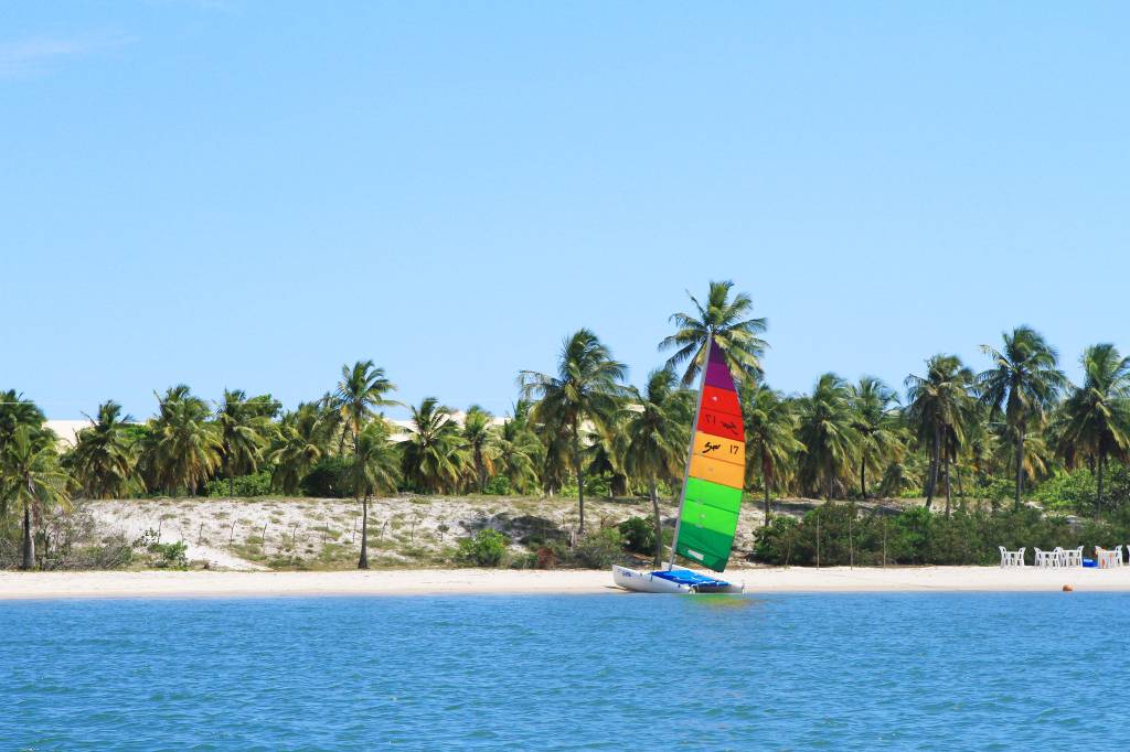 Vista da praia de Mangue Seco, no litoral sul da Bahia, durante o dia, com jangada de vela colorida atracada na areia e coqueiros e céu sem nuvens ao fundo