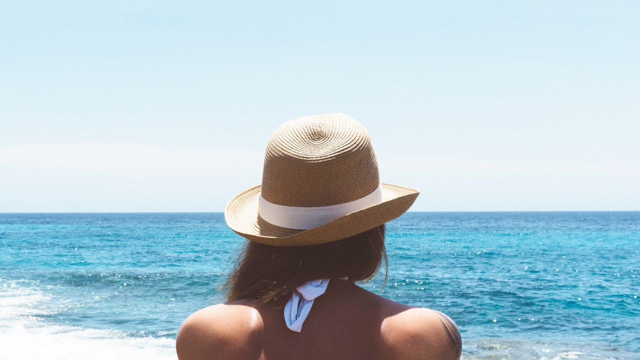 Mulher de chapéu, de costas, olha para o mar azul em praia