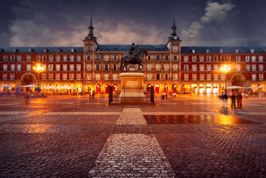 <strong>Plaza Mayor – <a href="https://viagemeturismo.abril.com.br/cidades/madri/" target="_blank" rel="noopener">Madri</a> – <a href="https://viagemeturismo.abril.com.br/paises/espanha/" target="_blank" rel="noopener">Espanha</a></strong> Boa parte das cidades espanholas e mesmo aquelas fundadas e conquistadas por eles nas Américas têm sua <strong>Plaza Mayor</strong>. Centro gravitacional da vida cívica, mercado e sede de edifícios governamentais, tudo girava ao seu redor. A mais bela e definitiva dessas praças não poderia estar em outro lugar, senão na capital do país. Ao longo de sua história, a Plaza Mayor de Madri foi palco de touradas, execuções e autos de fé da radical Inquisição espanhola, um local onde igreja e coroa atestavam seu poder perante o povo. O formato retangular e a relativa simetria no desenho dos edifícios é obra de sucessivos monarcas, mas boa parte das atuais características se devem a Filipe III, cuja estátua está fixada no centro da praça. Cafés e restaurantes espalham sob as arcadas centenas de mesas durante o dia, um ótimo lugar para ver a vida passar  <em>Busque hospedagem em Madri</em>