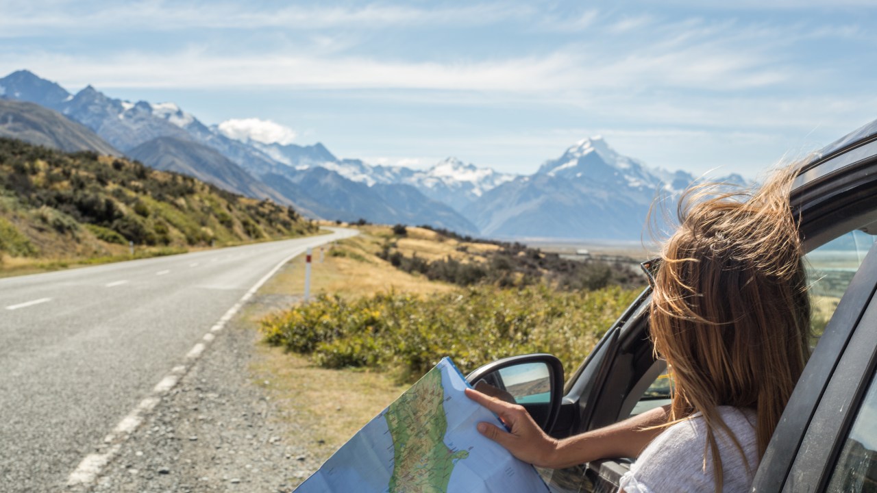 Mulher observa mapa em rodovia com cadeia de montanhas com picos nevados ao fundo