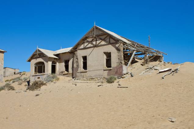 <strong>Cidade Fantasma de Kolmanskop, Namíbia</strong>    Os desertos africanos possuem um fascínio que desperta a curiosidade dos visitantes, sobretudo o Mar de Areia da Namíbia, tombado pela Unesco em 2013 como Patrimônio Mundial. Foi a areia, aliás, a responsável por engolir as casas construídas na cidade de Kolmanskop. No início de 1900, a região foi ocupada pelos alemães, que viram nela a chance de enriquecer graças à exploração de diamantes. O excesso da atividade, no entanto, fez com que os minérios sofressem um forte esgotamento. Na década de 1950, o abandono por parte de seus moradores fez com que a constante limpeza da areia insistente que ocupava as suas ruas fosse completamente abandonada. Hoje, o governo mantém a região habitável para que os turistas curiosos que passeiam por ali possam continuar a explorar a área