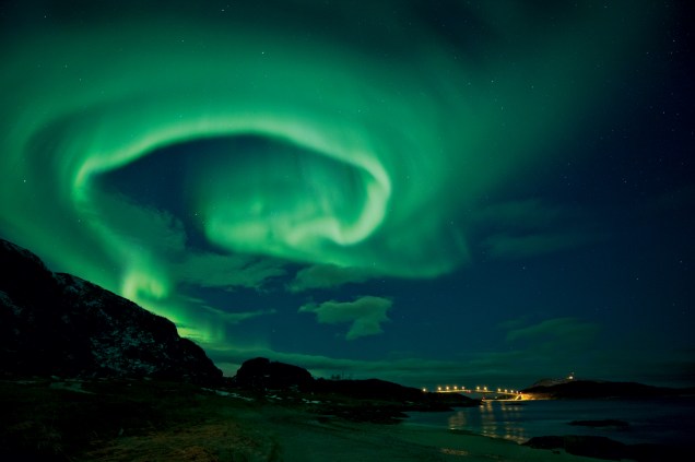 Uma aurora paira sobre a ponte Sommarøy, na ilha Kvaløy, no norte da Noruega, em uma semana de atividade solar intensa. As auroras são causadas pelo impacto de partículas carregadas nos gasesda atmosfera. Mais comuns perto dos polos, as auroras também ocorremem latitudes mais baixas durante tempestades solares fortes.