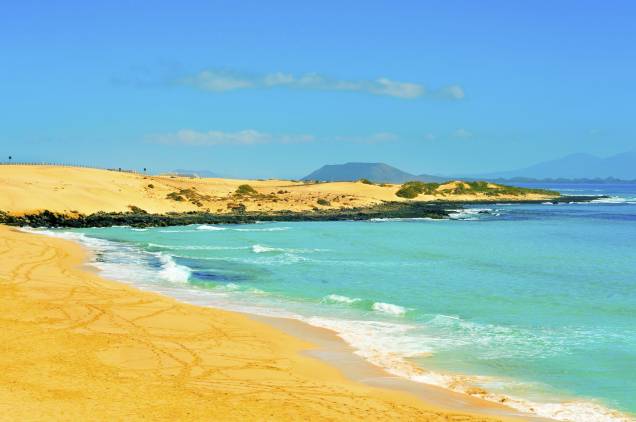 <strong>Fuerteventura, Ilhas Canárias, <a href="http://viajeaqui.abril.com.br/paises/espanha" rel="Espanha" target="_self">Espanha</a> </strong>                “Ilha da tranquilidade”: é assim que muitos conhecem Furteventura, marcada por praias tranquilas em meio a cenários rurais. Localizadas em uma região vulcânica, muitas de suas praias podem apresentar areia mais escura. Um dos grandes destaques são as <strong>Dunas de Corralejo</strong>, localizadas em uma área de preservação ambiental e acopladas a um parque natural de rara beleza                <em><a href="http://www.booking.com/region/es/canary-islands.pt-br.html?sid=5b28d827ef00573fdd3b49a282e323ef;dcid=1/?aid=332455&label=viagemabril-as-mais-belas-praias-do-mediterraneo" rel="Veja preços de hotéis próximos a Fuerteventura no Booking.com" target="_blank">Veja preços de hotéis próximos a Fuerteventura no Booking.com</a></em>