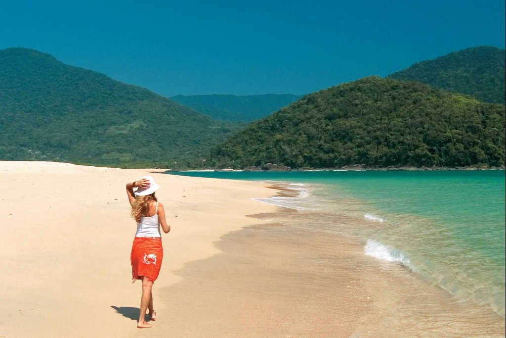 Mulher de férias caminha descalça na areia de uma praia, com montanhas cobertas de árvores ao fundo