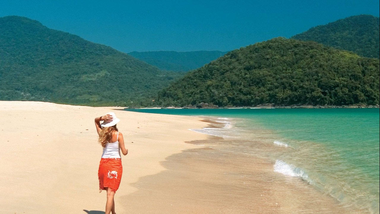 Mulher de férias caminha descalça na areia de uma praia, com montanhas cobertas de árvores ao fundo