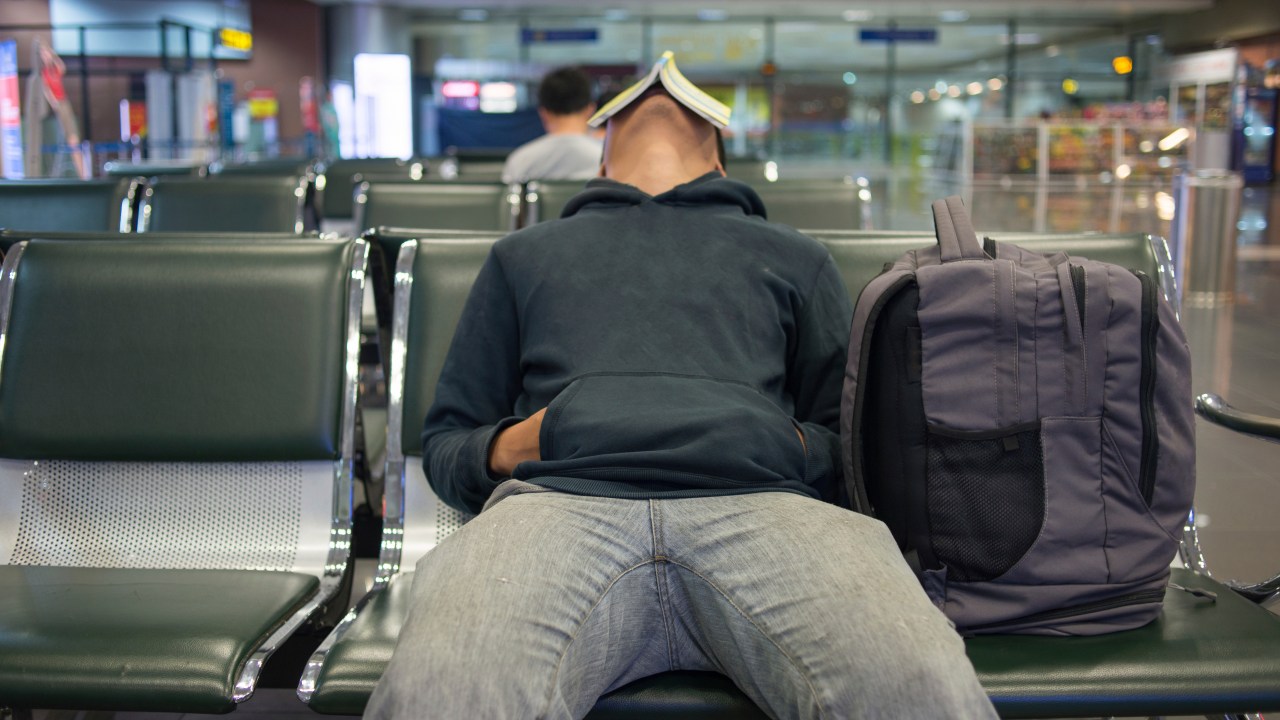 Homem espera em sala de embarque de aeroporto com livro sobre a cara