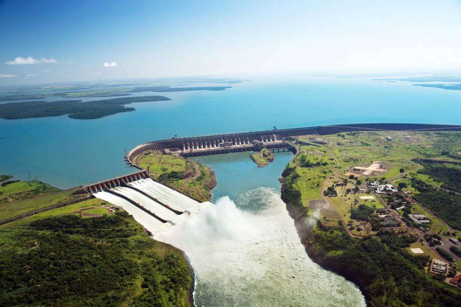 A imagem mostra a vista aérea de uma grande usina hidrelétrica. As comportas estão abertas então é possível ver água abundante no vertedouro
