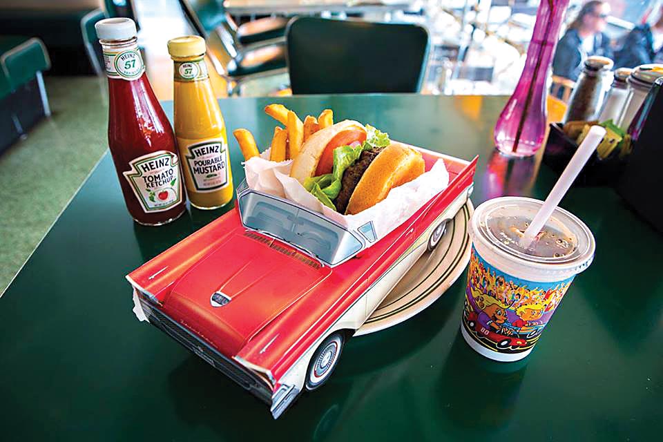 Um hambúrguer e batatas fritas se encontram dentro de um minicarro conversível, que funciona como um prato em cima de uma mesa. Do lado direito, um copo de plástico guarda um refrigerante e, à esquerda, há dois potes: um de ketchup e outro de mostarda