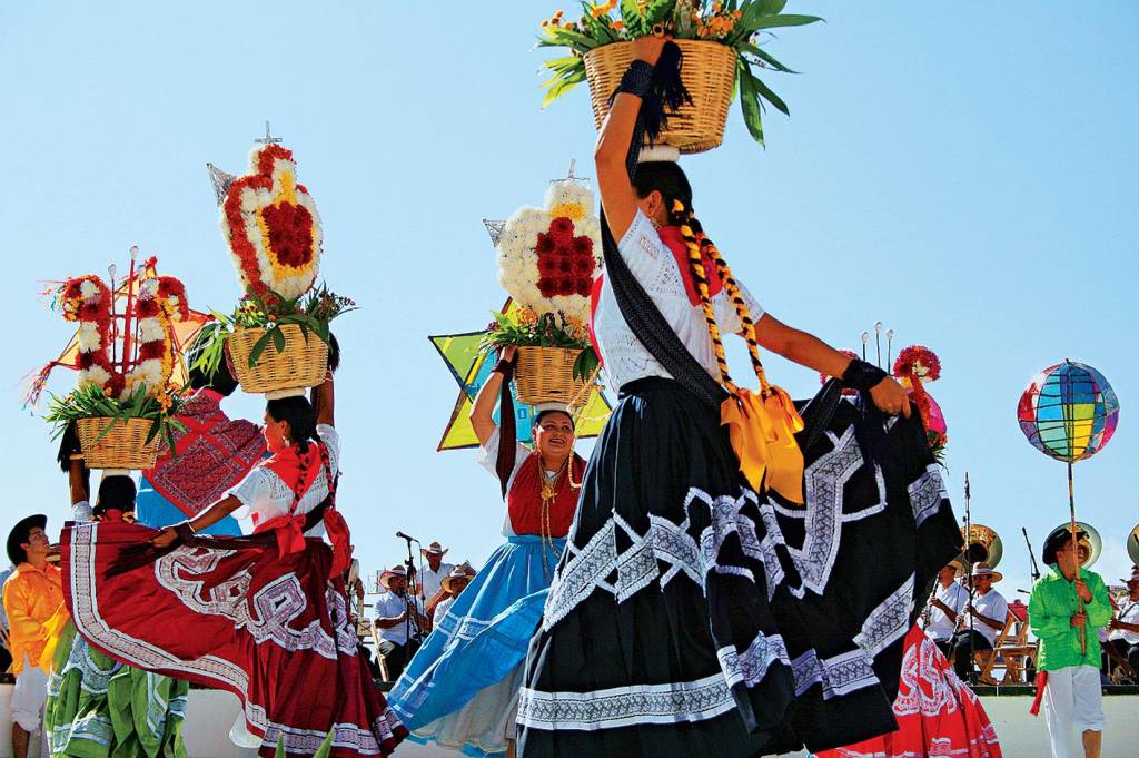 Mulheres dançam ao som de um grupo musical ao vivo, com longas tranças e saias compridas. Em uma mão, seguram uma cesta de flores apoiada na cabeça, enquanto com a outra levantam uma ponta da saia para dançar