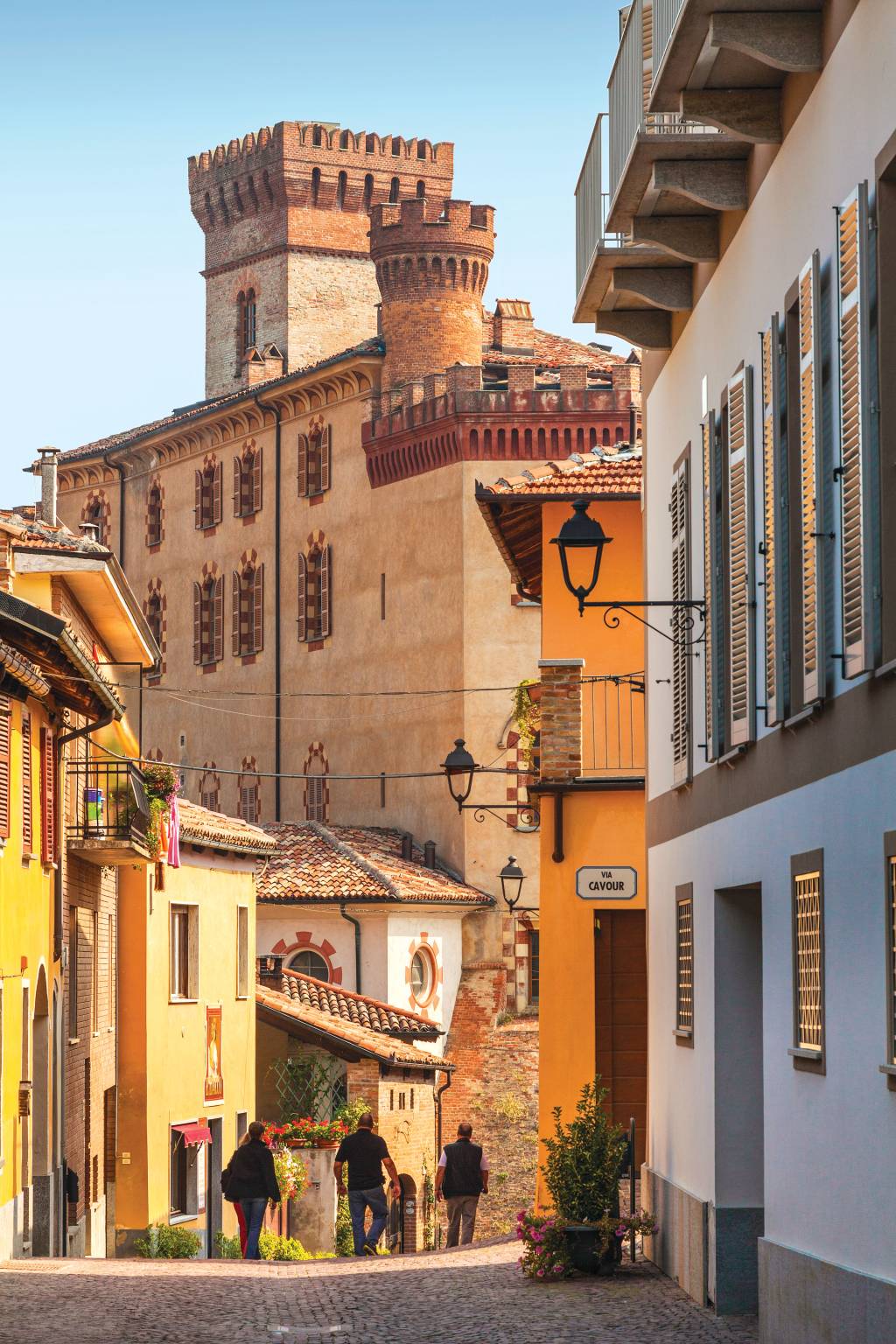 Três pessoas de costas andam em uma rua estreita e cheia de casinhas. Ao fundo, uma das seções do Castelo em Grinzane Cavour