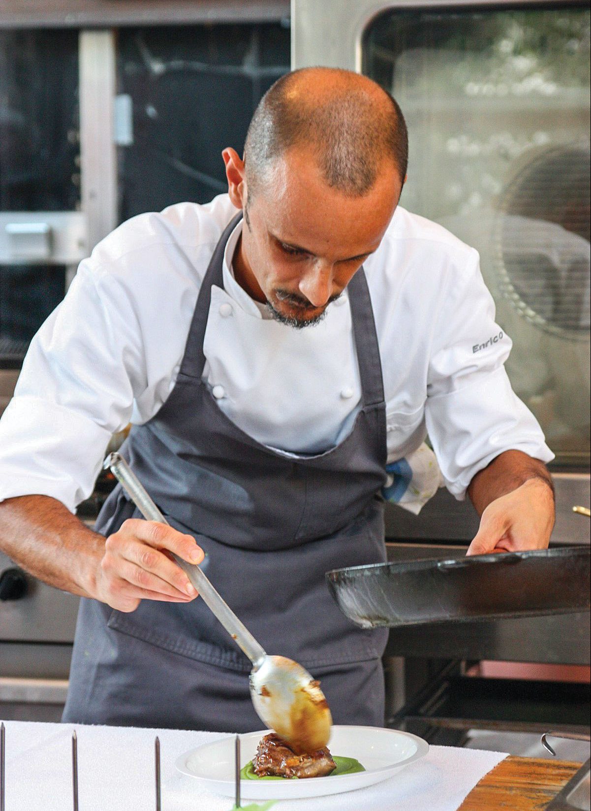 Chef Enrico Crippa, vestindo um avental, com uma panela na mão e uma colher na outra, espalha molho sobre o pedaço de uma carne