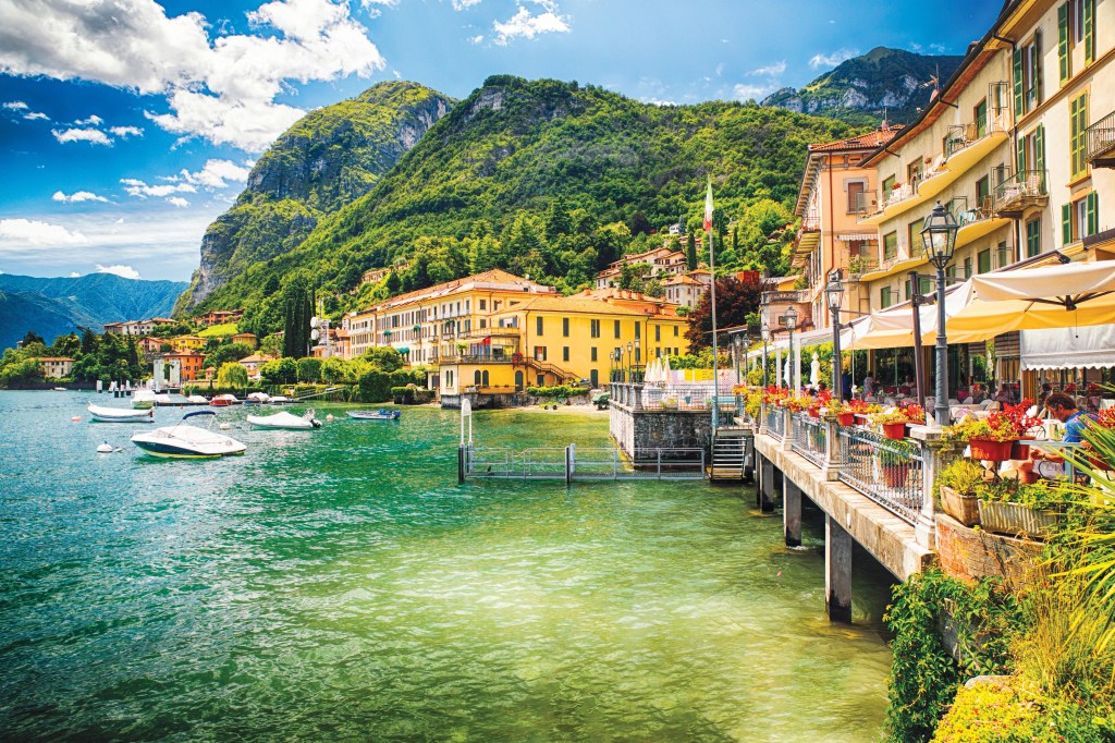 Do lado direito, um restaurante charmoso e à beira d'água é decorado com flores. Ao fundo, casarões amarelos. À esquerda, lanchas estão ancoradas no Lago Como