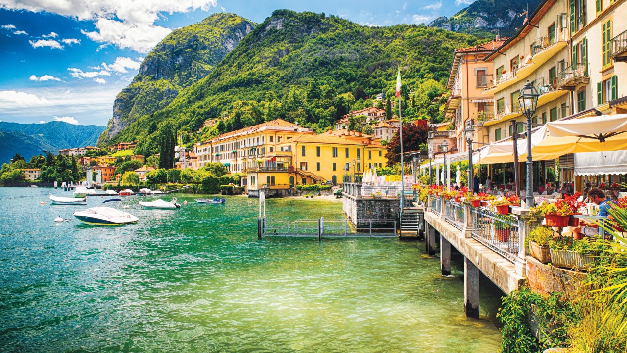 Do lado direito, um restaurante charmoso e à beira d'água é decorado com flores. Ao fundo, casarões amarelos. À esquerda, lanchas estão ancoradas no Lago Como