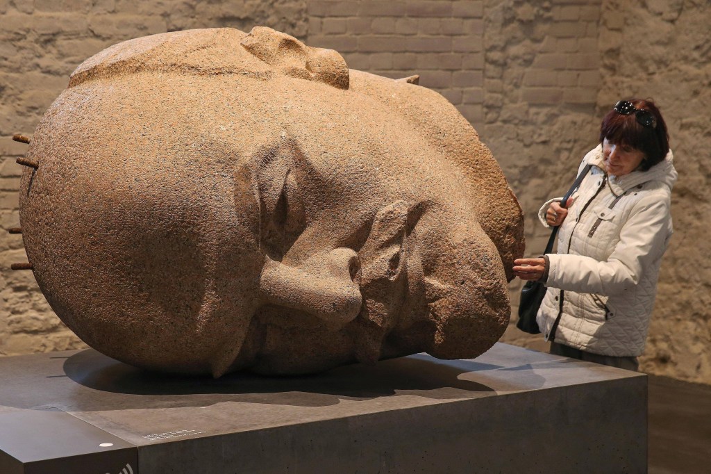 Um busto enorme de pedra com feições detalhadas do revolucionário Lênin. Ao lado, uma mulher toca na escultura, que tem o dobro de seu tamanho.