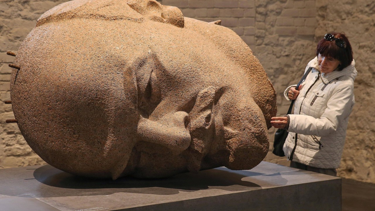 Um busto enorme de pedra com feições detalhadas do revolucionário Lênin. Ao lado, uma mulher toca na escultura, que tem o dobro de seu tamanho.