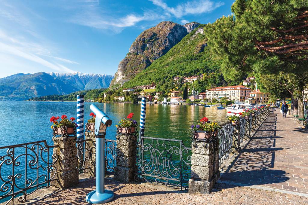 Uma luneta que aponta para o horizonte do Lago Como decora o canto de uma calçada de pedras, que é ladeada por árvores e com uma grade repleta de florzinhas