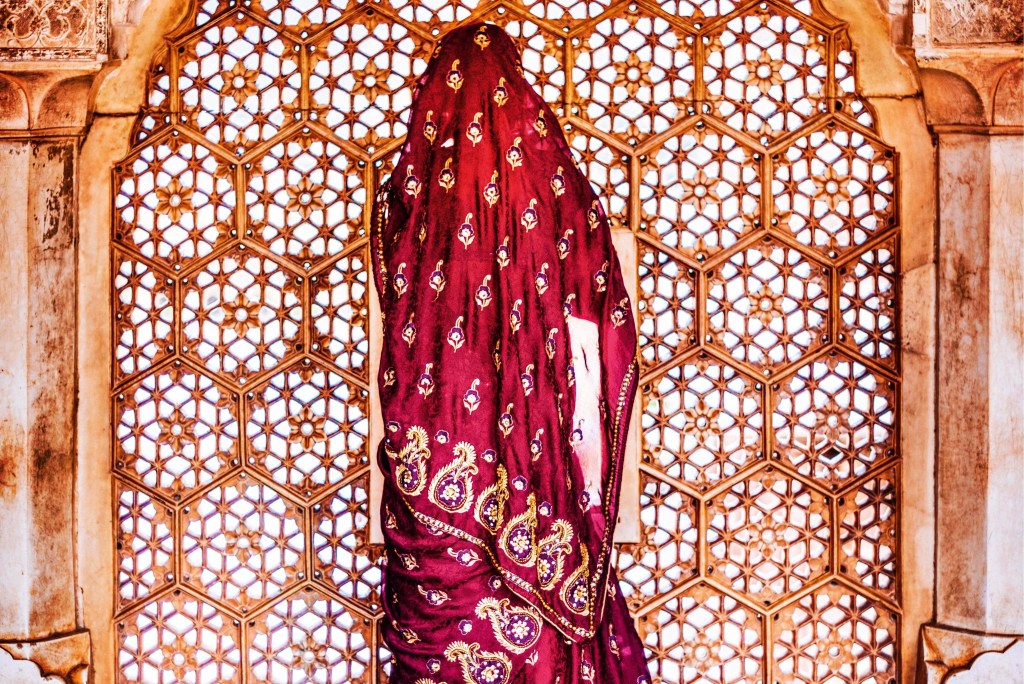 Mulher vestida em um sári se posiciona de frente a uma janela, cuja abertura está divida em furos formados por várias mandalas esculpidas