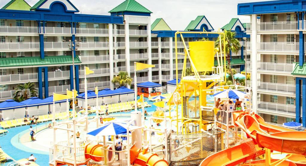 Um pequeno parque aquático, com crianças descendo escorregadores e em piscinas. Ao fundo, prédios com as varandas e os quartos dos hóspedes