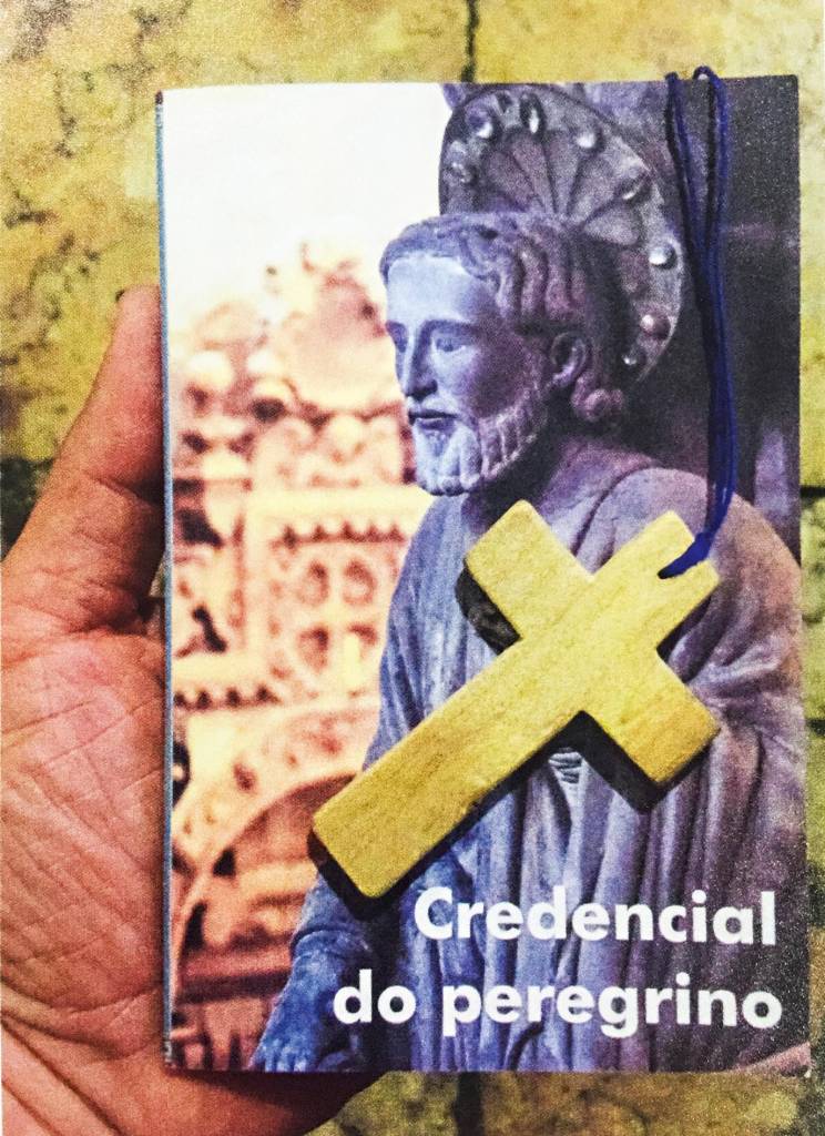 Uma mão segura um livreto chamado "credencial do peregrino", cuja capa é a foto de uma estátua de um santo. Uma cruz de madeira está posicionada em cima da capa.