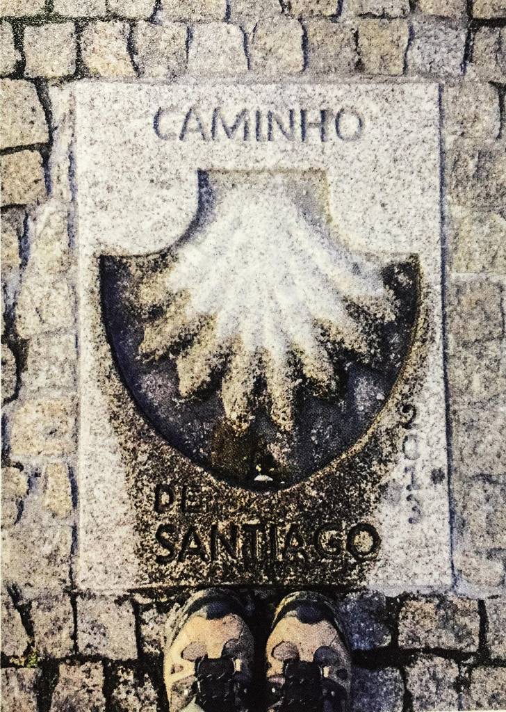 Uma concha esculpida no chão, com a palavra "Caminho" acima e "de Santiago" abaixo. Em frente à concha estão duas botas, com os bicos apontando em sua direção.