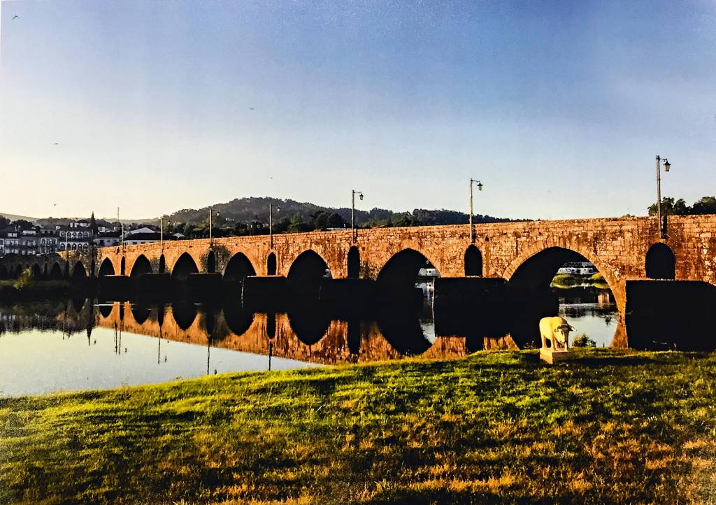 Ponte de Lima, Santiago de Compostela, Espanha Dividindo a imagem em dois, a ponte de Lima, formada por diversos arcos com bases submersas, parece se aproximar do leitor graças ao ângulo lateral de fotografia. Do lado de cá, grama; do lado de lá, alguns prédios antigos.