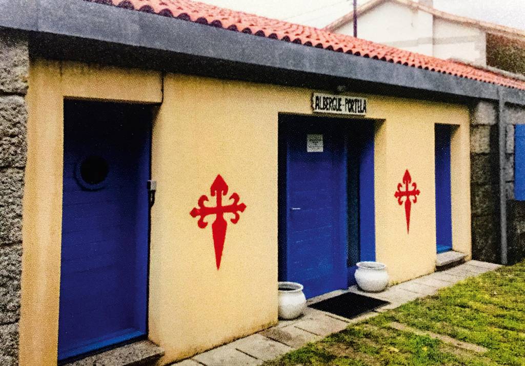 Três portas vistas de um ângulo lateral. Sobre a porta central, uma placa com os dizeres "Albergue-Portela" está posicionada, e há dois brasões pintados nos muros entre as portas.