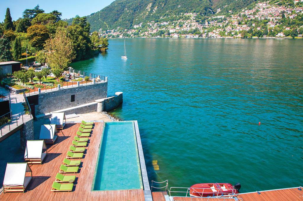 Panorâmica de parte do Lago Como. Em primeiro plano e à beira d'água, cadeiras reclináveis e a piscina compõem o limite do Il Sereno Hotel. Ao fundo, casinhas amontoam-se sob os pés das montanhas