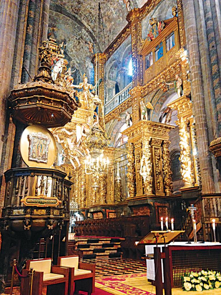Interior de uma catedral, com tetos altos, pisos de ladrilho de diferentes padrões e grupos de pequenas colunas suportando a estrutura, assim como pinturas e estátuas de santos e madonas, grande parte em ouro.