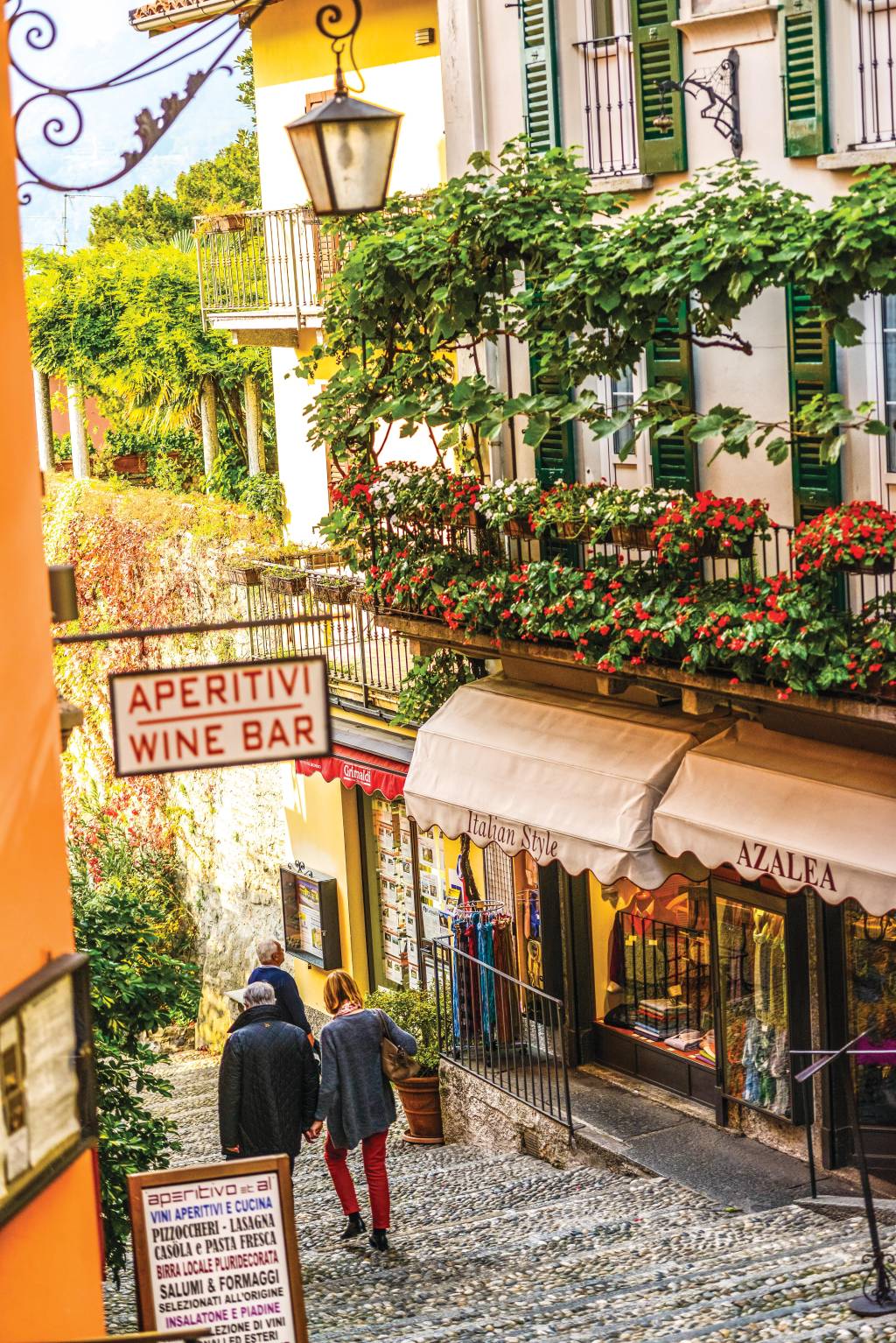 Rua em Bellagio, Lago Como, Itália Um casal desce as escadarias de pedra de uma estreita ladeira. Os prédios que a ladeiam são altos e charmosos, com lojinhas, varandinhas cheias de flores e lâmpadas de rua antigas. Do lado esquerdo, uma placa indica: