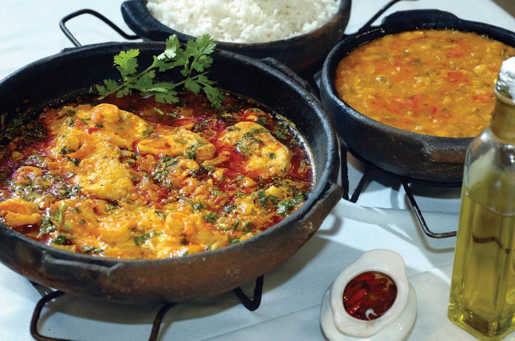 Moqueca dentro de um panela de barro, ao lado de outras que servem arroz e molho de camarão. À esquerda, azeite e um potinho com pimenta capixaba