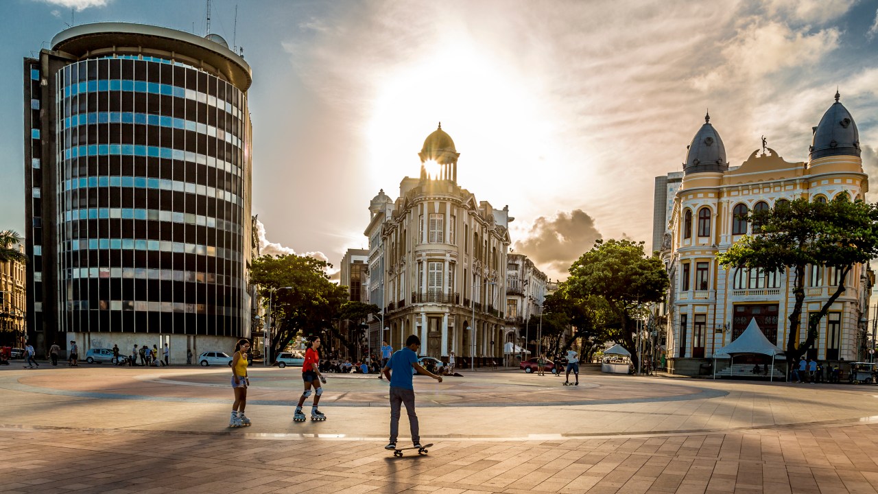 Pôr do sol na praça do Marco Zero no Recife Antigo, pessoas passeiam no calçadão colorido com vista para prédios históricos ao fundo