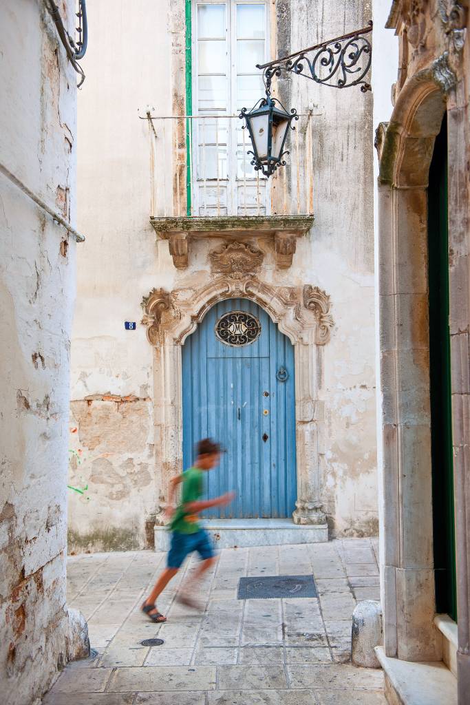 Uma rua com uma grande e ricamente decorada porta. Um menino pequeno corre em frente ao lugar