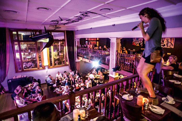 Um cantora está em cima de uma mesa, no segundo andar de um restaurante, se apresentando para a plateia de clientes do restaurante