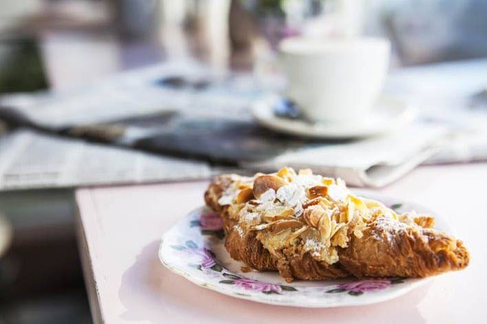 Um croissant com cobertura de amêndoas está em cima de um pequeno prato, com florezinhas. Em cima da mesma mesa, mas ao fundo da foto, uma xícara situa-se em cima de vários jornais