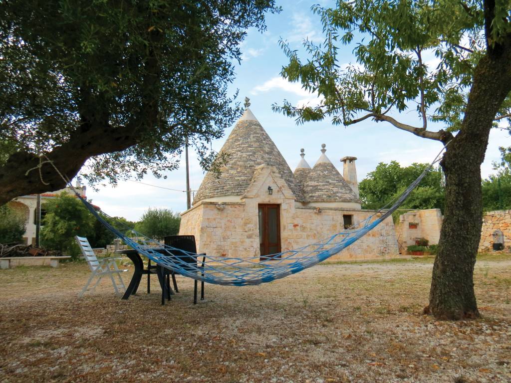 Casa em estilo trullo, com um quintal na frente, no qual duas árvores suportam uma rede