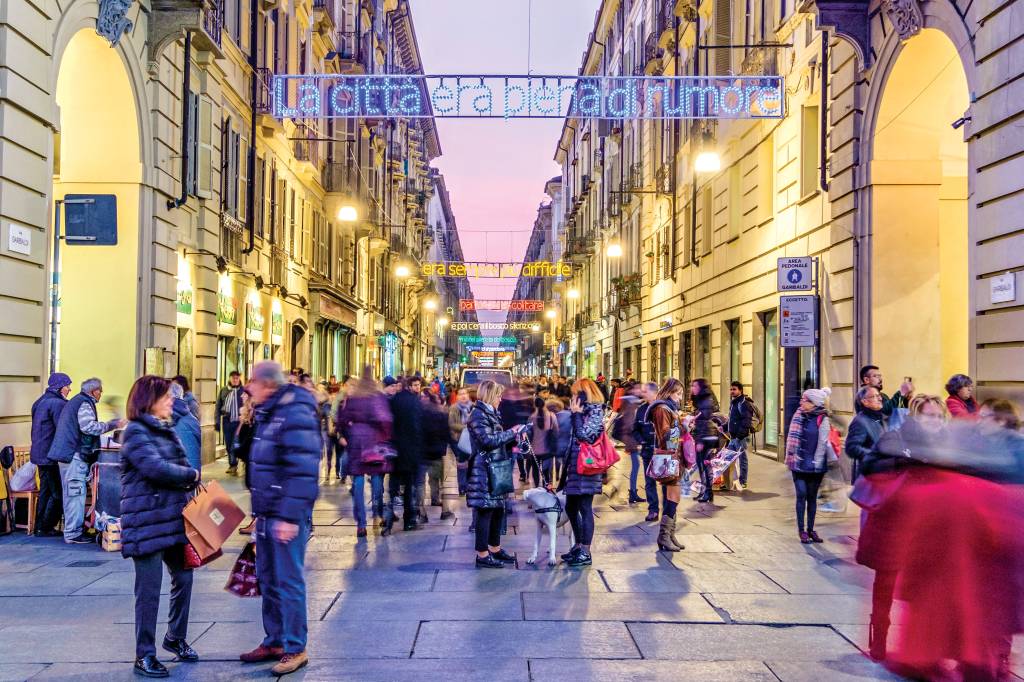 Pessoas andam, conversam e interagem no meio da Via Garibaldi, uma rua estreita e com prédios altos, na cidade de Turim