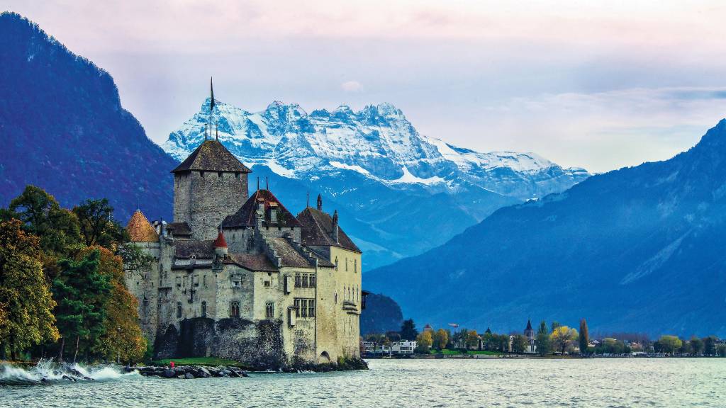 Um castelo se impõe à beira de um lago, cercado por árvores e com os Alpes visíveis ao fundo