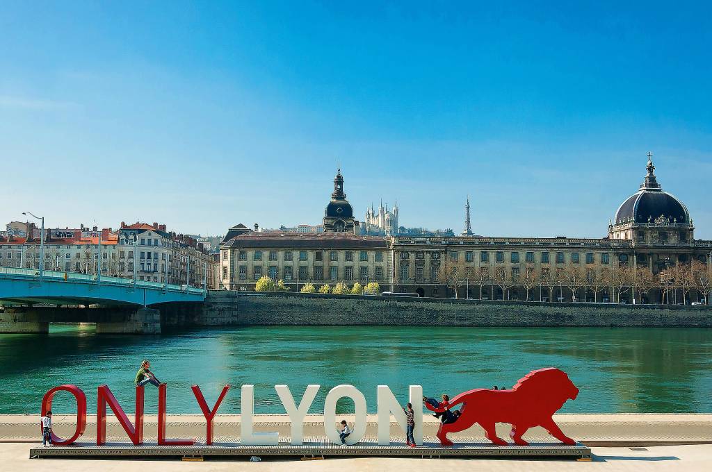 Um letreiro enorme com os dizeres "Only Lyon" e um leão esculpido na frente de um rio e de prédios franceses, no fundo. Crianças e turistas sobem nas letras gigantes e no leão.