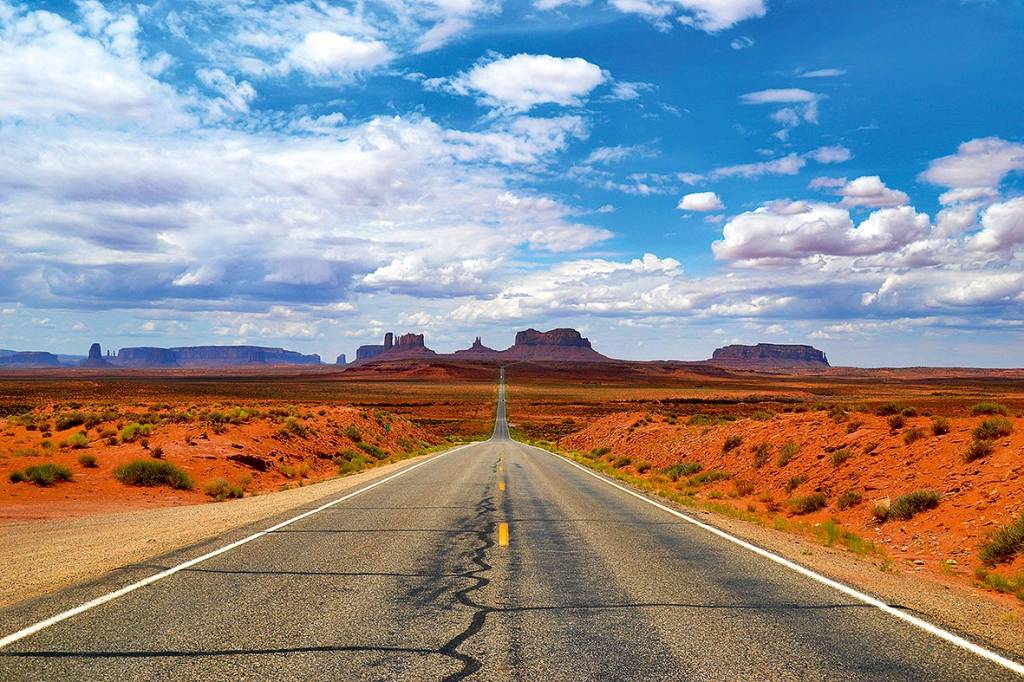 Uma estrada vazia corre para o horizonte, onde há vários cânions. Ao lado, paisagem desértica típica dos Estados Unidos.