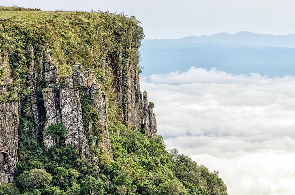 Um precipício com várias plantas e árvores ao longo do lado da montanha. Ao fundo, várias nuvens e, no horizonte, uma cadeira de montanhas. A foto é vista, provavelmente, de outro ponto da Serra do Rio do Rastro.