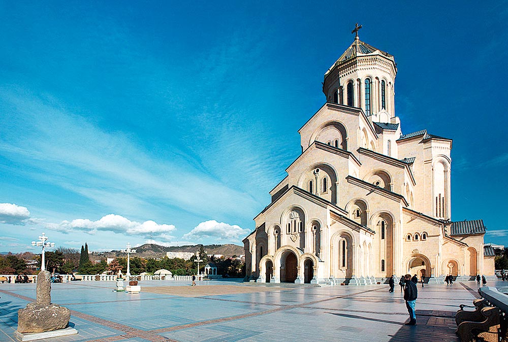 Uma grande catedral, com uma arquitetura que divide a sua altura em camadas, localizada em uma grande praça com montanhas ao fundo