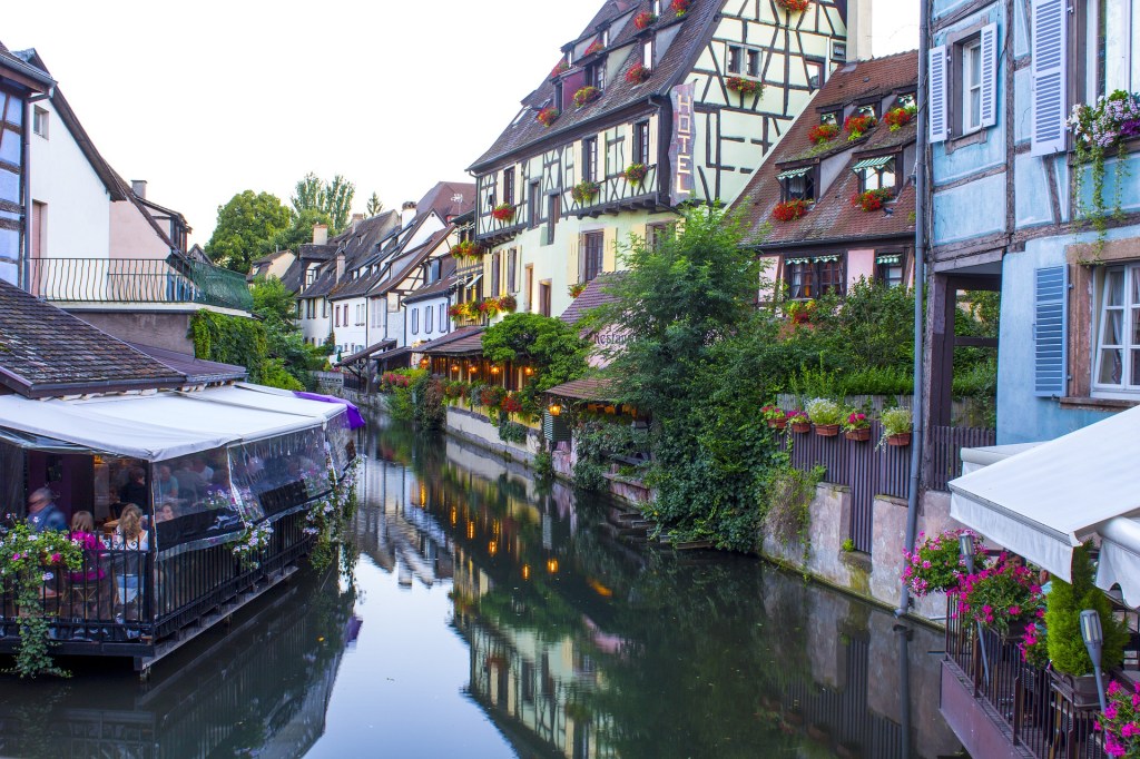 Visão do curso de um rio, com casas de estilo típico francês em uma margem e um restaurante na outra