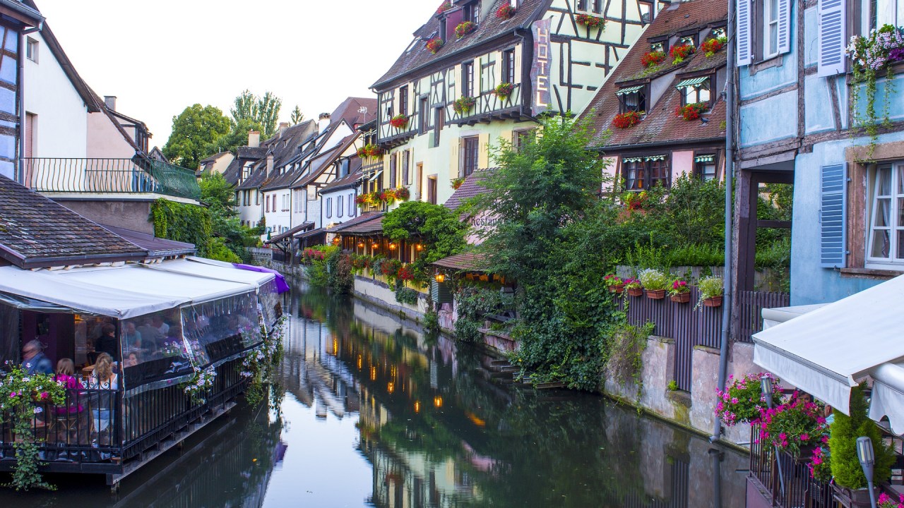 Visão do curso de um rio, com casas de estilo típico francês em uma margem e um restaurante na outra