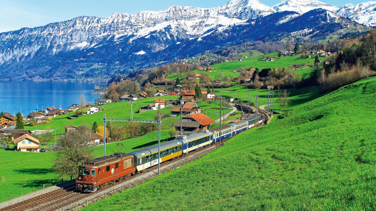 Um trem atravessa uma pequeno vilarejo, as casinhas espalhadas pelo campo de grama e esparsos grupos de árvores, com a cadeia alpina nos fundos