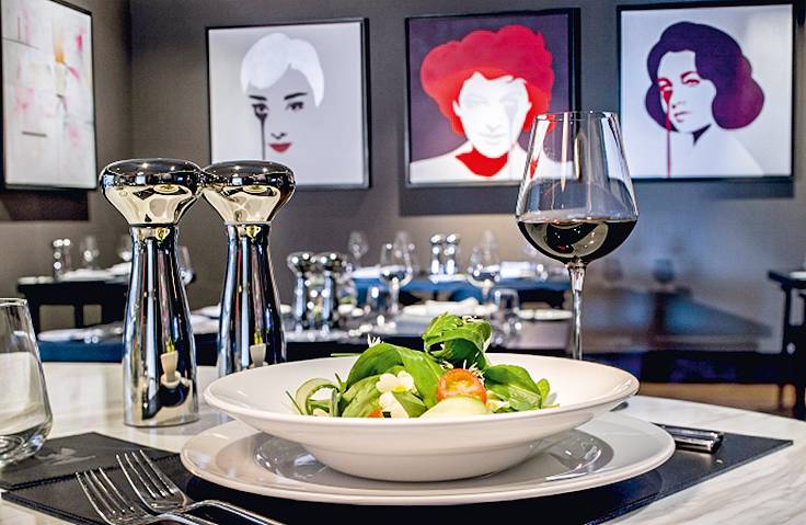 Prato de salada em restaurante requintado, cercado por talheres lustrosos e quadros minimalistas de mulheres