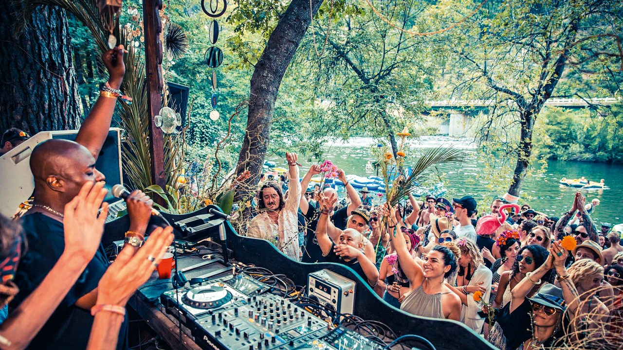 DJ organiza festa à beira de um rio, apinhando pessoas em frente à mesa com acessórios extravagantes