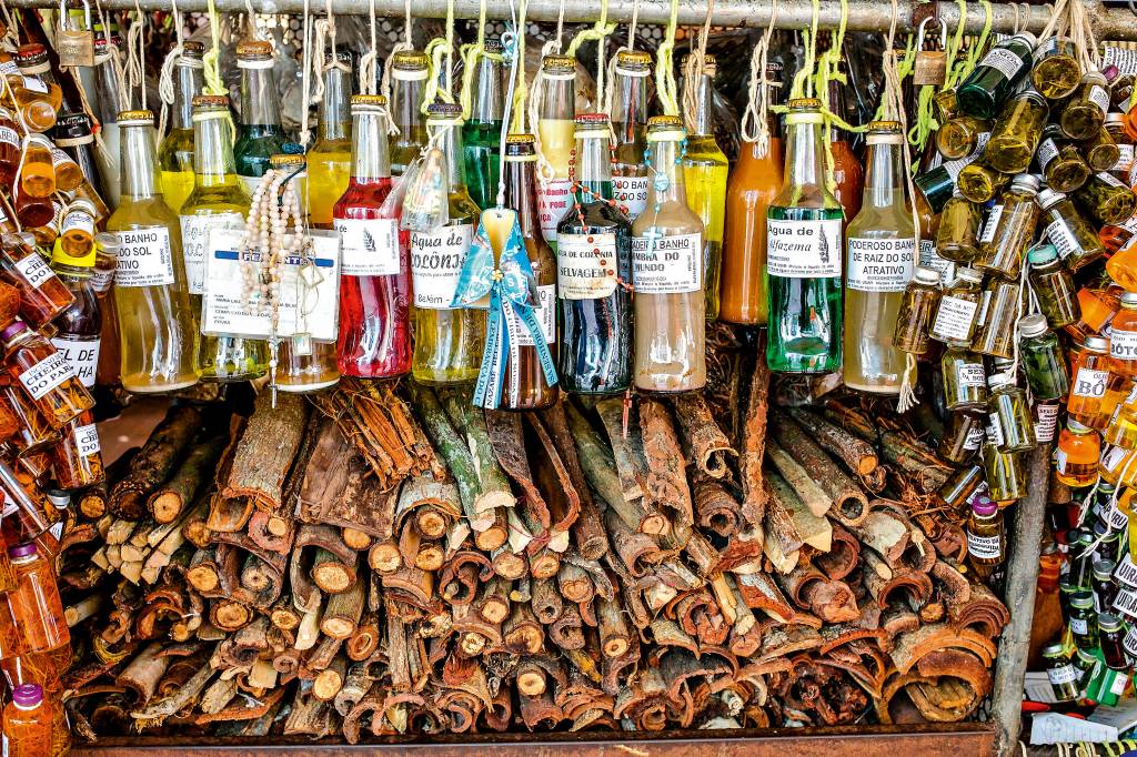 Garrafas de vidro penduradas por cordas amarradas ao pescoço vendem diversos tipos de líquidos. dos lados, garrafinhas também estão à venda, penduradas ao longo estande, e embaixo há uma pilha de cascas de árvore