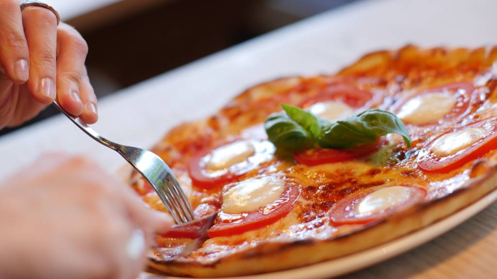 Pizza Tonda, Itália A imagem é a foto de uma pessoa comendo individualmente uma pizza de formato redondo. É possível perceber que a pizza leva queijo, tomates e manjericão.